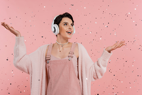 amazed and happy woman in wireless headphones standing under confetti on pink background