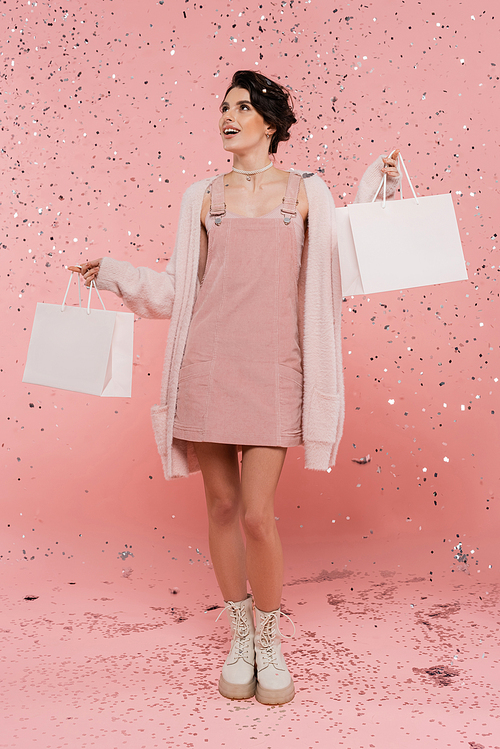 full length of happy and trendy woman standing with shopping bags under confetti on pink