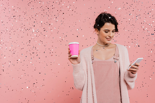 trendy brunette woman with coffee to go messaging on smartphone under confetti on pink background