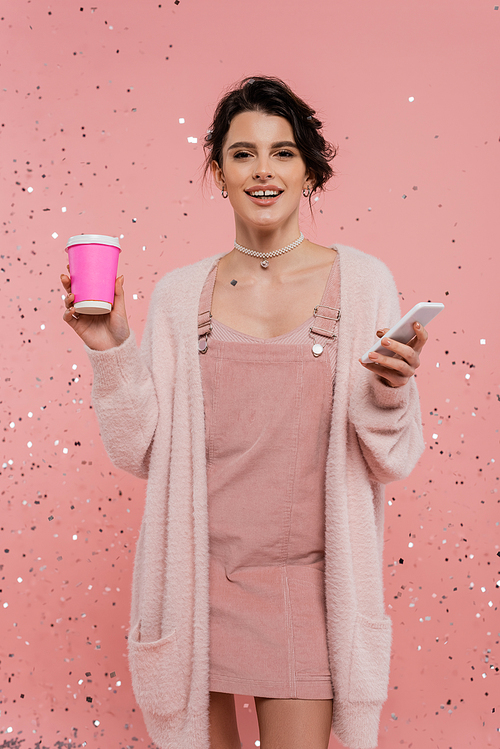 happy brunette woman with paper cup and mobile phone looking at camera on pink background