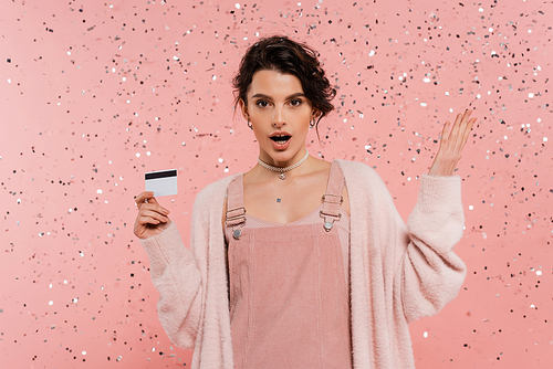 amazed woman in strap dress and cardigan holding credit card and pointing with hand under confetti on pink background