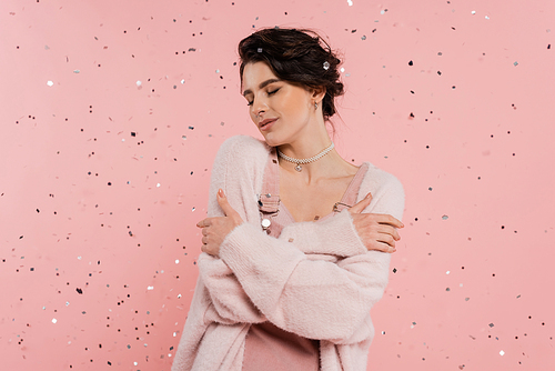 brunette woman in cozy cardigan embracing herself while standing with closed eyes under confetti on pink background
