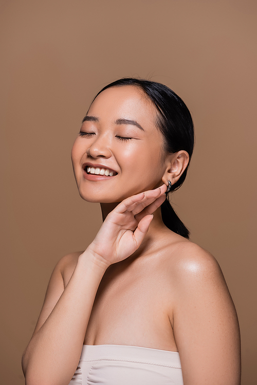 Happy asian woman in top closing eyes and touching face isolated on brown