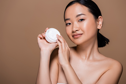 Pretty asian woman with naked shoulders holding container with cosmetic cream isolated on brown