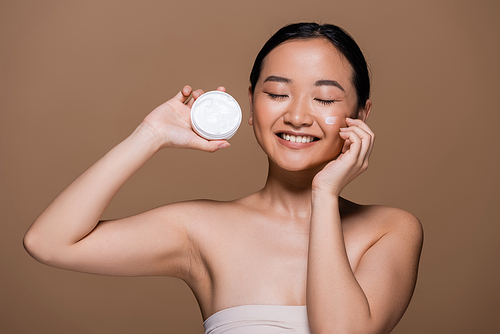 Positive asian woman with naked shoulders holding face cream isolated on brown