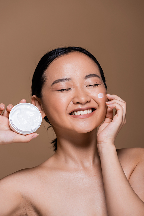 Pretty asian woman with naked shoulders holding cosmetic cream isolated on brown