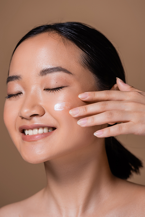 Smiling asian woman applying face cream isolated on brown