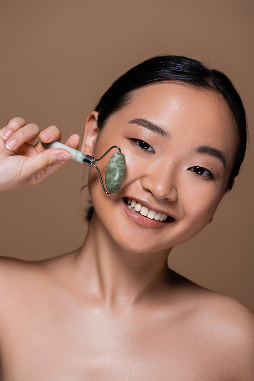 Cheerful asian woman with naked shoulders holding jade roller isolated on brown