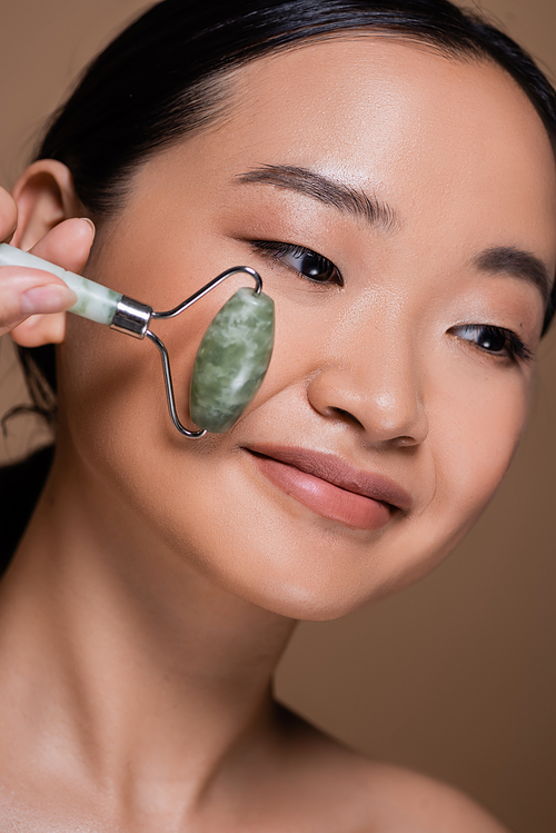 Portrait of asian woman holding jade roller near cheek isolated on brown