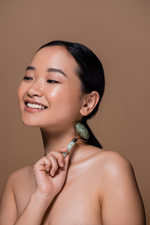 Smiling young asian woman with naked shoulders holding jade roller isolated on brown