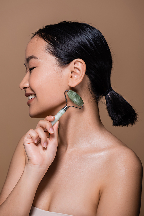 Side view of smiling asian woman with naked shoulders holding jade roller isolated on brown