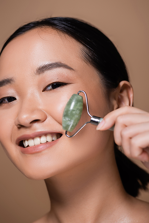 Portrait of cheerful asian woman holding jade roller and looking at camera isolated on brown