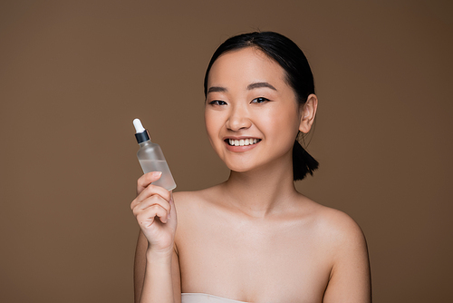 Positive asian woman with naked shoulders holding bottle of essential oil isolated on brown