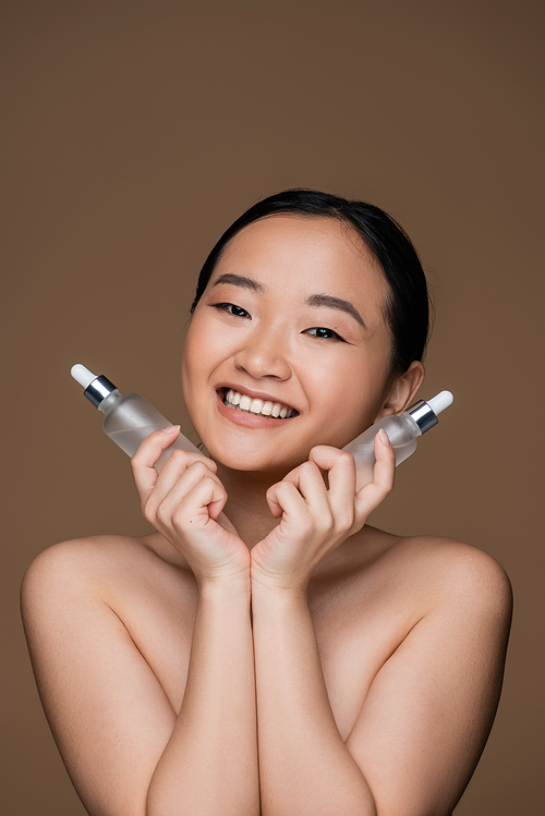 Cheerful asian woman with naked shoulders holding cosmetic serums isolated on brown