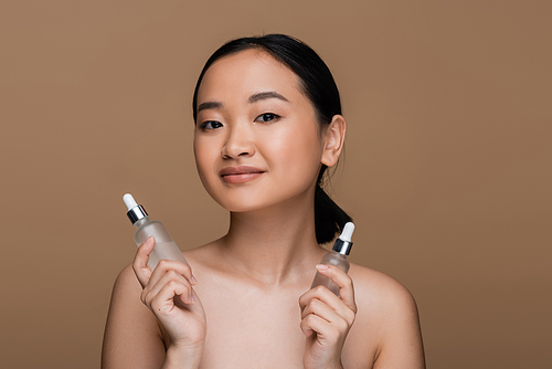 Pretty asian model with naked shoulders holding cosmetic serums isolated on brown