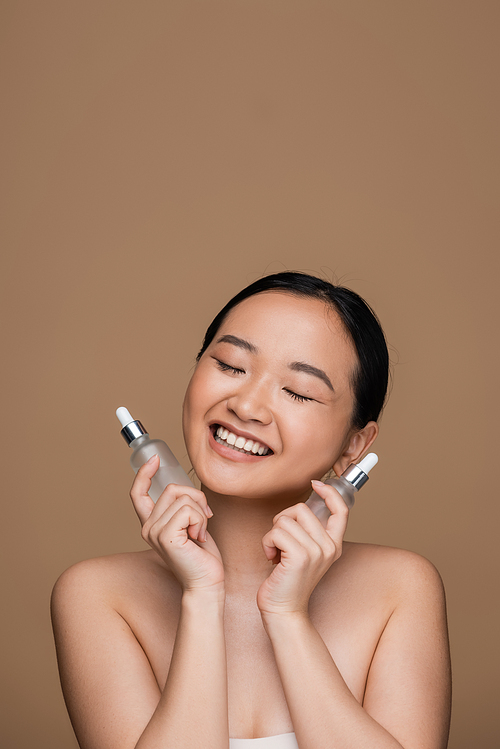Pleased asian woman holding cosmetic serums isolated on brown