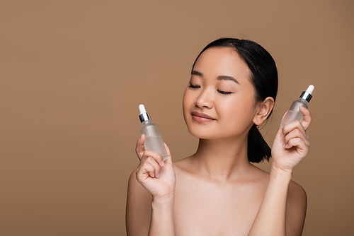 Pretty asian woman with naked shoulders holding bottles of serums isolated on brown