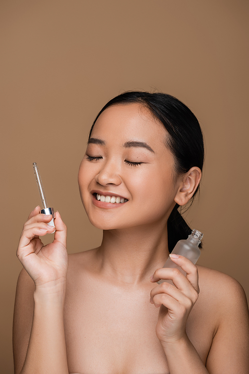 Positive asian woman with naked shoulders holding serum isolated on brown