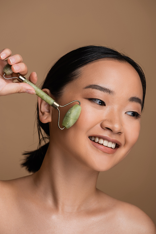 Positive asian model with naked shoulders using jade roller on face isolated on brown