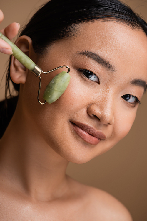 Portrait of brunette asian woman using jade roller on face isolated on brown