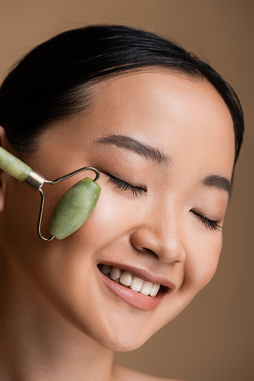 Pleased asian woman using jade roller on cheek isolated on brown