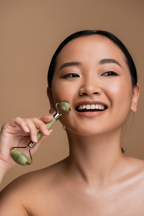 Positive asian model with naked shoulder using jade roller isolated on brown