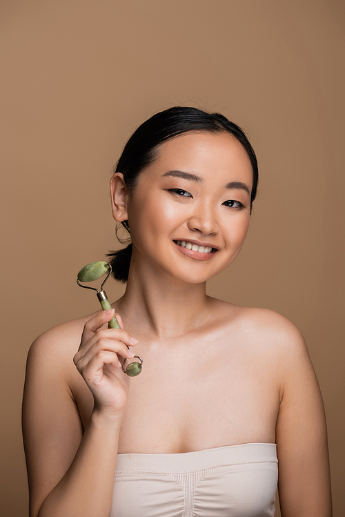 Positive asian model in beige top holding jade roller and looking at camera isolated on brown
