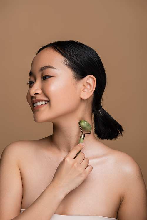 Happy asian woman with naked shoulders using jade roller on neck isolated on brown