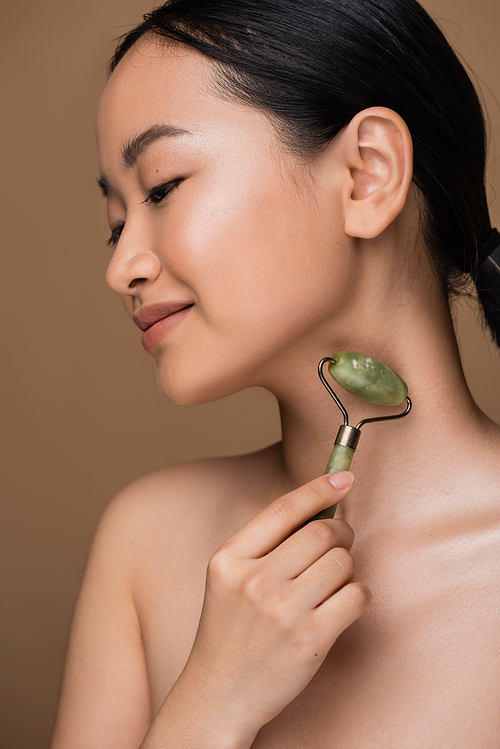 Asian woman with naked shoulder using jade roller on neck isolated on brown