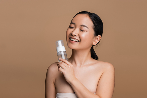 Positive asian model with naked shoulders holding cleansing foam isolated on brown