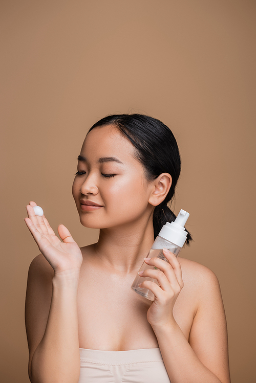 Young asian woman in top looking at cleansing foam on hand isolated on brown