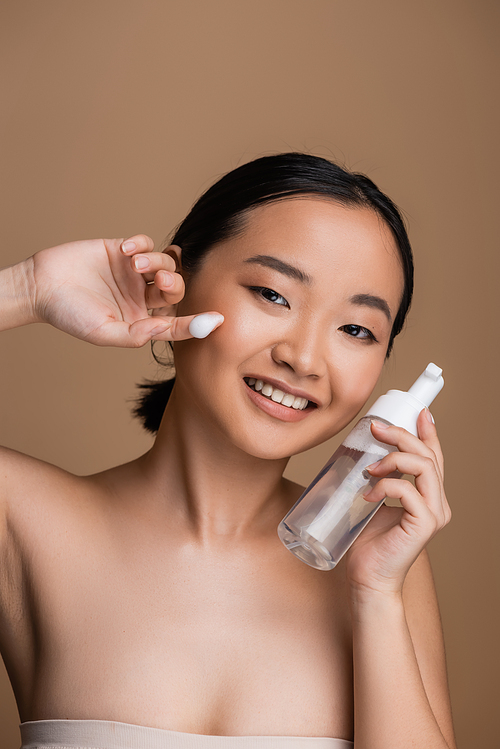 Positive asian model with naked shoulders holding cleanser with dispenser isolated on brown