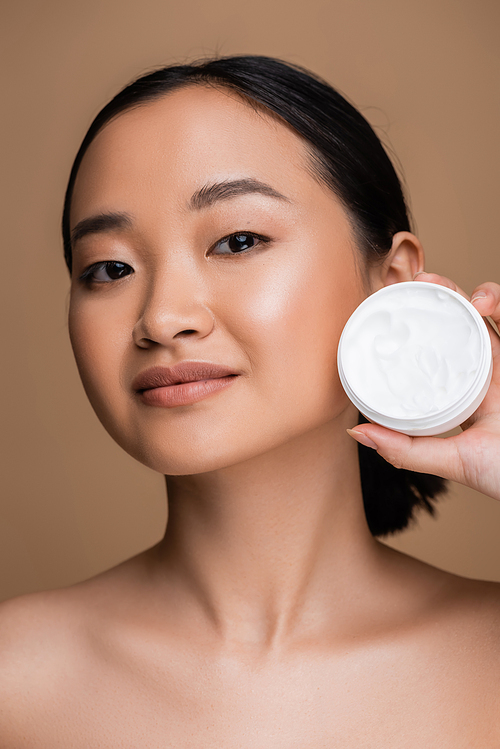 Pretty asian woman with naked shoulders and perfect skin holding cosmetic cream isolated on brown