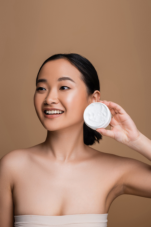 Cheerful asian model in top holding cosmetic cream isolated on brown
