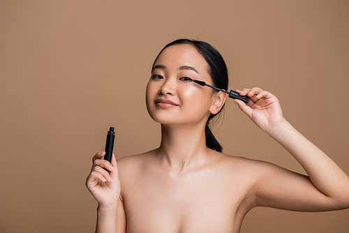 Young asian woman with naked shoulders holding mascara isolated on brown