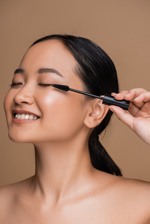 Cheerful asian woman with naked shoulders holding mascara applicator isolated on brown