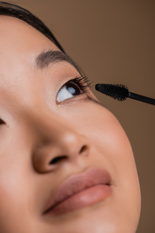Close up view of asian woman applying mascara isolated on brown