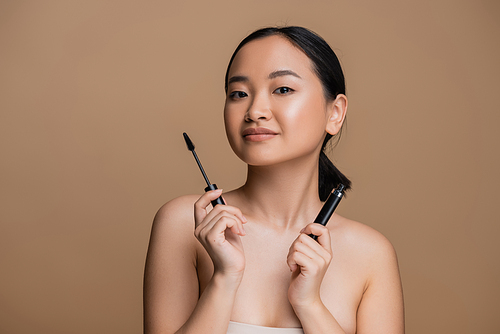 Pretty asian woman holding mascara and looking at camera isolated on brown