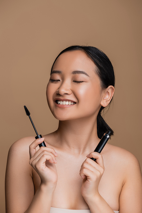 Cheerful asian woman with naked shoulders holding mascara isolated on brown