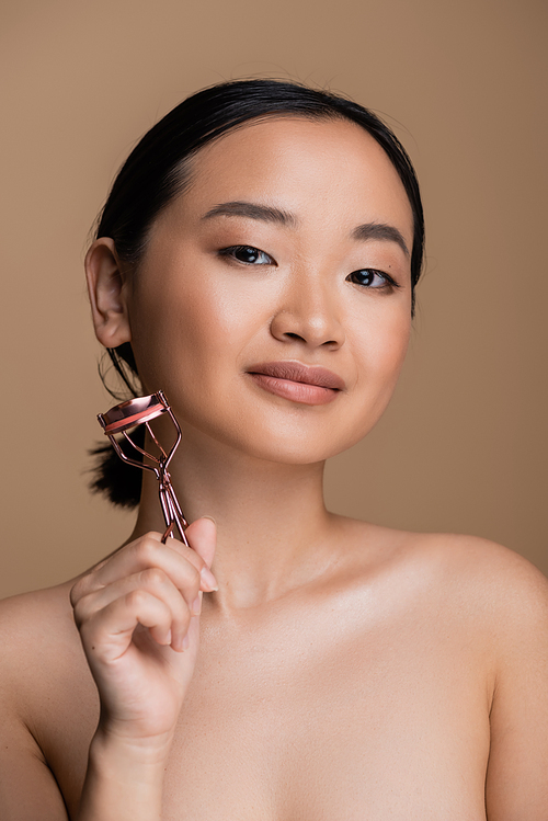 Pretty asian woman with naked shoulders holding lush curler isolated on brown