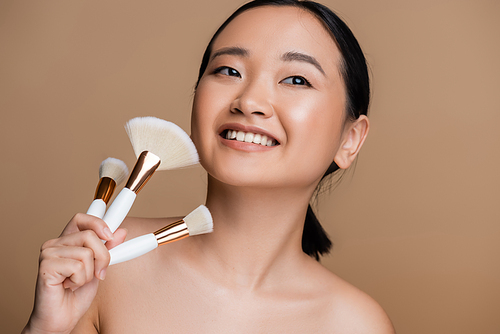 Pretty asian woman holding cosmetic brushes and looking away isolated on brown