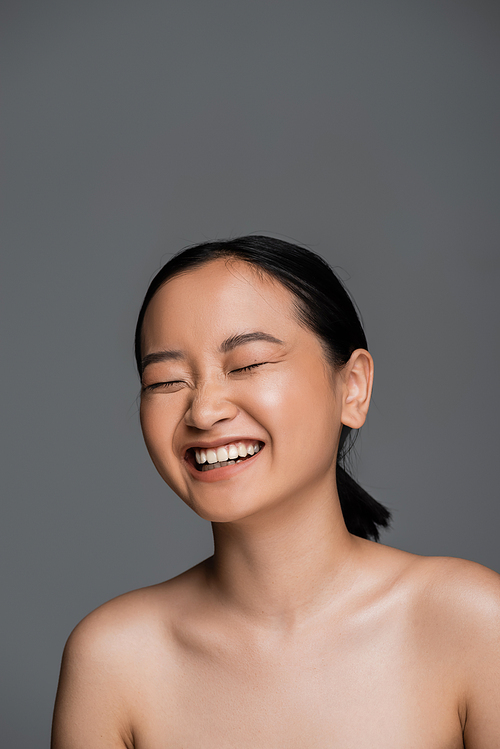 Cheerful asian woman with naked shoulders laughing isolated on grey