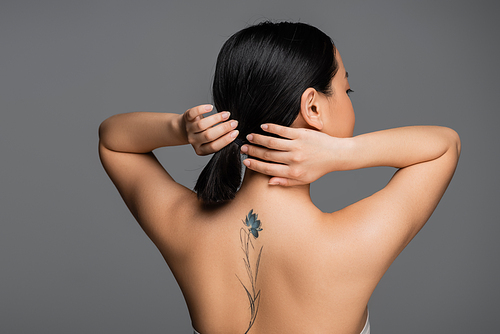 Brunette asian woman with tattoo on back touching ponytail isolated on grey