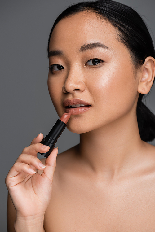 Portrait of young asian woman with naked shoulder holding lipstick isolated on grey