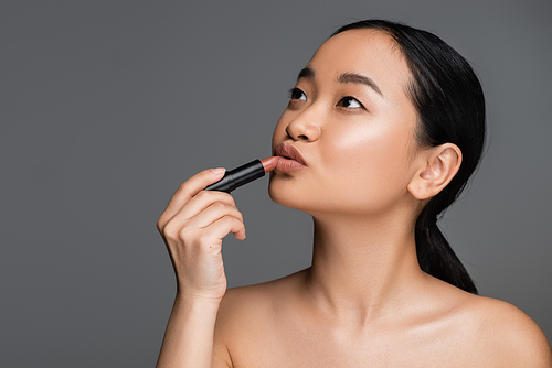 Pretty asian woman applying lipstick and looking away isolated on grey