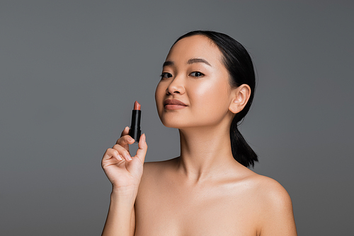 Pretty asian woman with naked shoulders holding beige lipstick isolated on grey