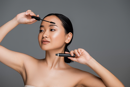 brunette asian woman with naked shoulders and perfect skin holding black mascara isolated on grey