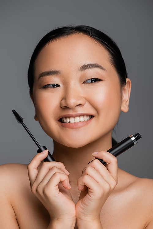happy asian woman with bare shoulders and natural makeup holding mascara isolated on grey