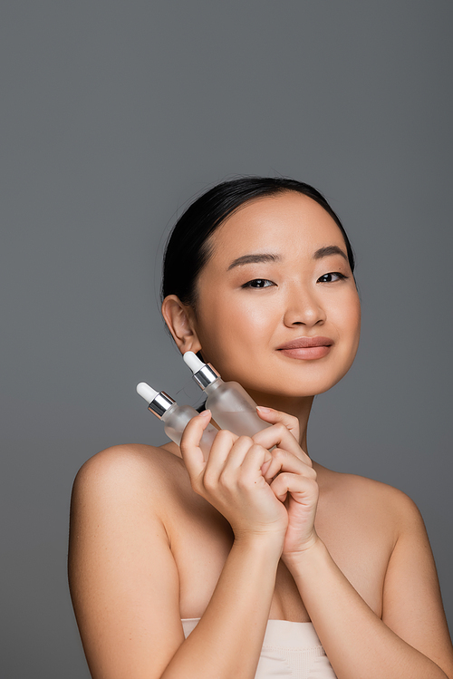 brunette asian woman with perfect skin holding bottles of natural cosmetic serum isolated on grey