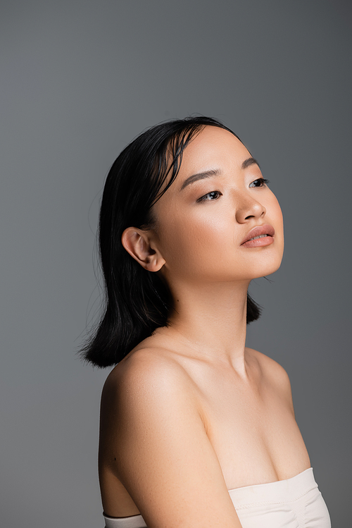 pretty asian woman with bare shoulders and nude makeup looking away isolated on grey